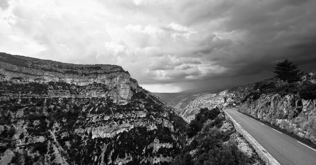 Route sinueuse de gorges, photographie noir et blanc montrant la route taillée dans la falaise calcaire, surplombant la vallée sous un ciel nuageux dramatique