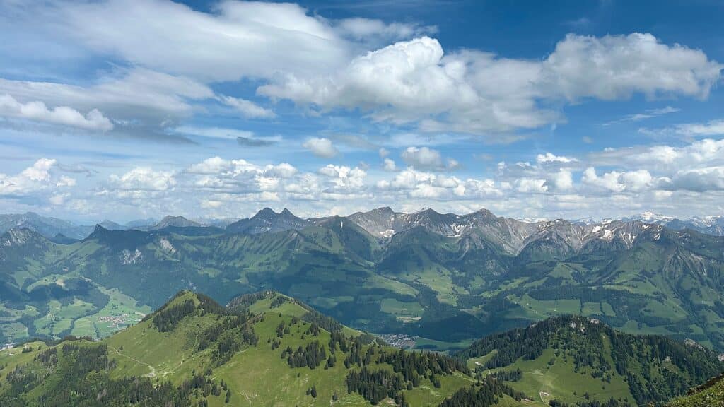 Panorama de montagnes verdoyantes sous un ciel bleu parsemé de nuages, sommets enneigés et vallées alpines en été.