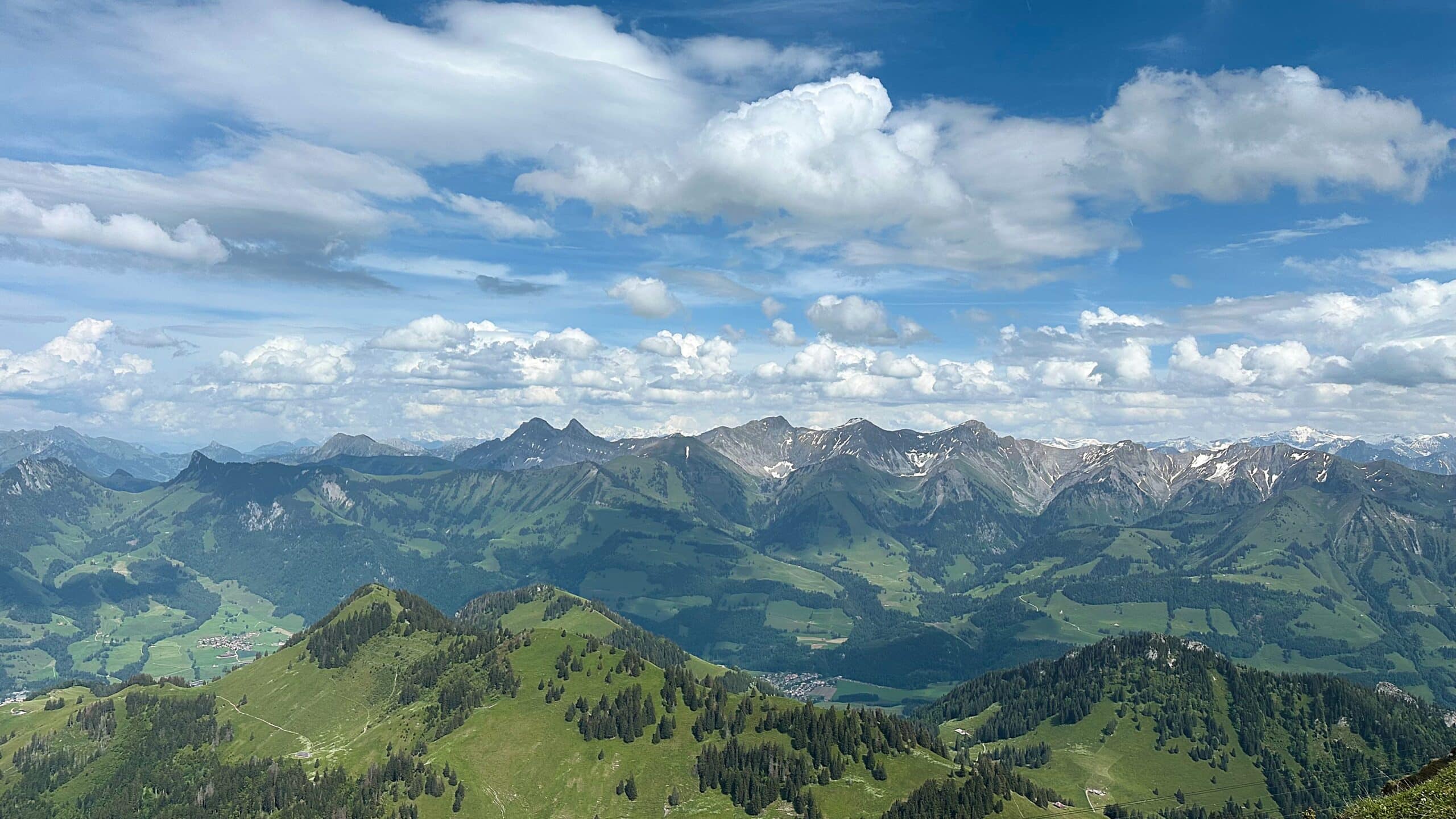 Panorama de montagnes verdoyantes sous un ciel bleu parsemé de nuages, sommets enneigés et vallées alpines en été.
