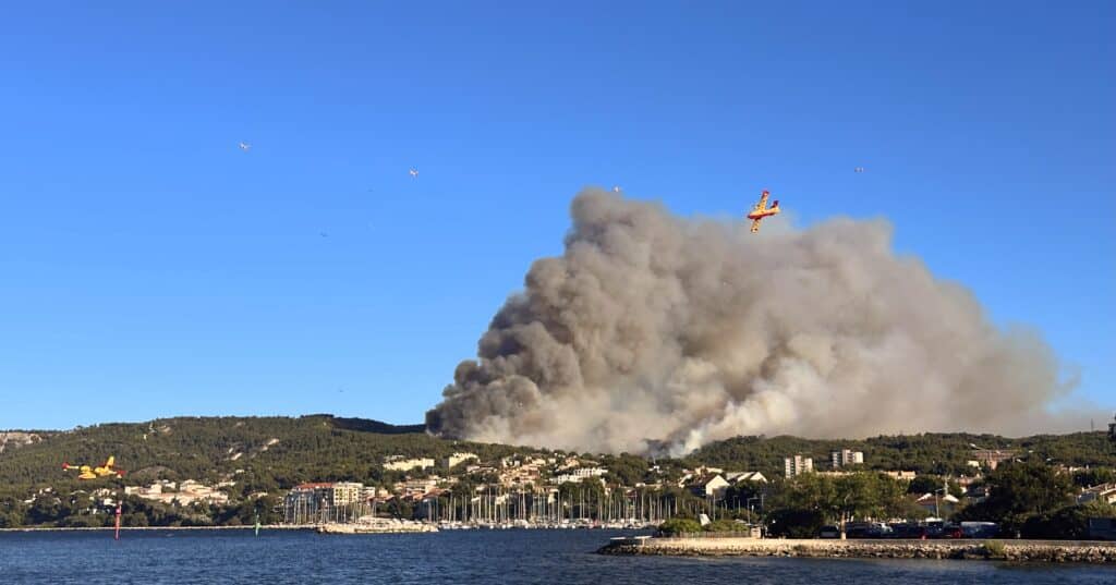 Plusieurs Canadair et oiseaux survolent une ville côtière avec épaisse fumée d'incendie, port et bâtiments visibles.