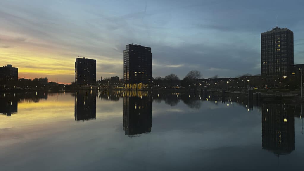 Immeubles se reflétant dans un canal au crépuscule, ciel bleu et jaune, lumières urbaines allumées et eau calme créant un miroir parfait.