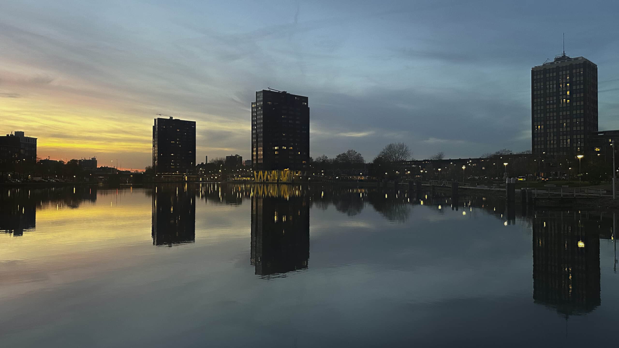 Immeubles se reflétant dans un canal au crépuscule, ciel bleu et jaune, lumières urbaines allumées et eau calme créant un miroir parfait.