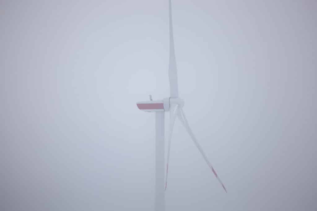 Éolienne blanche avec pales aux extrémités rouges, isolée dans un brouillard blanc épais, ambiance minimaliste et brumeuse.