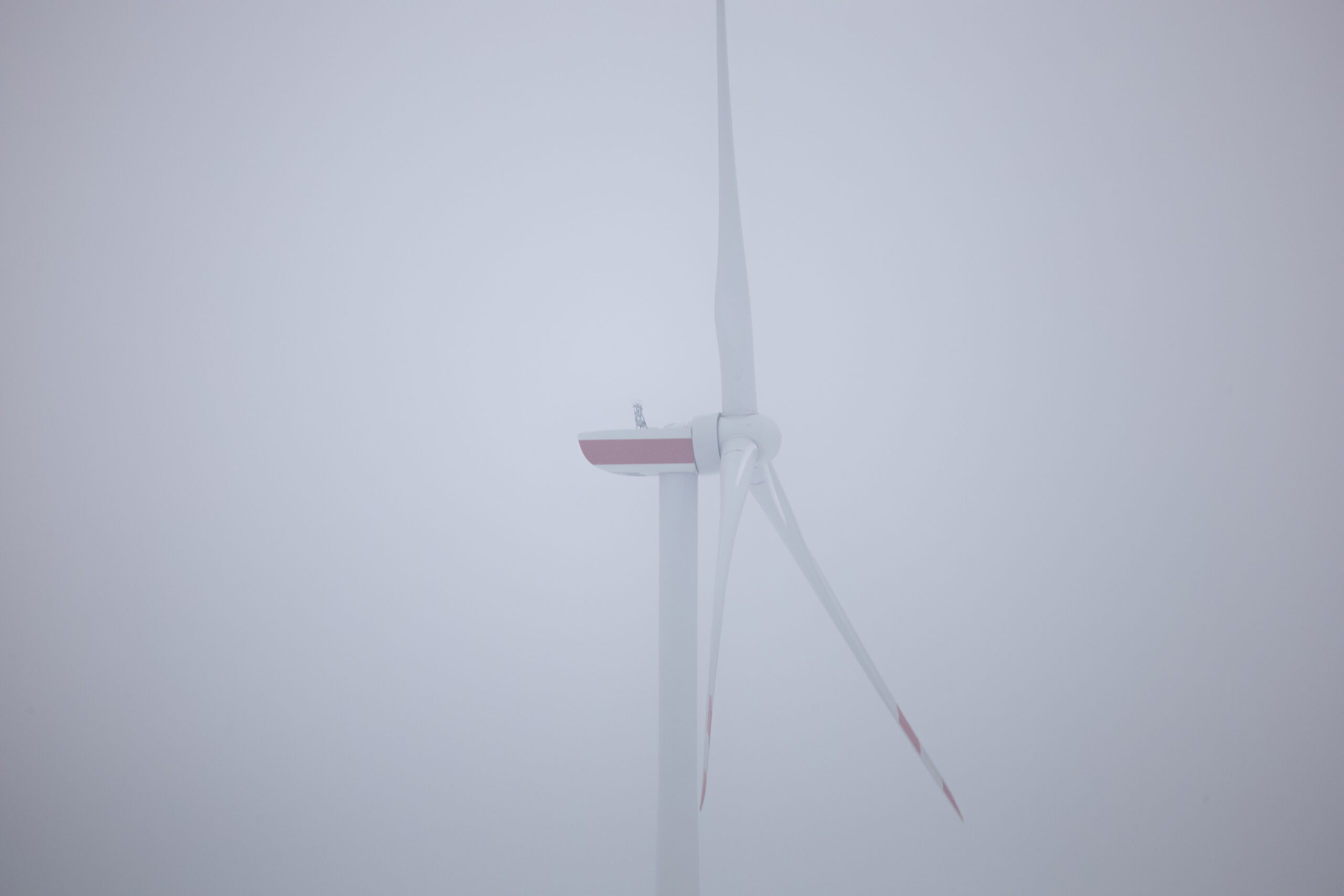 Éolienne blanche avec pales aux extrémités rouges, isolée dans un brouillard blanc épais, ambiance minimaliste et brumeuse.