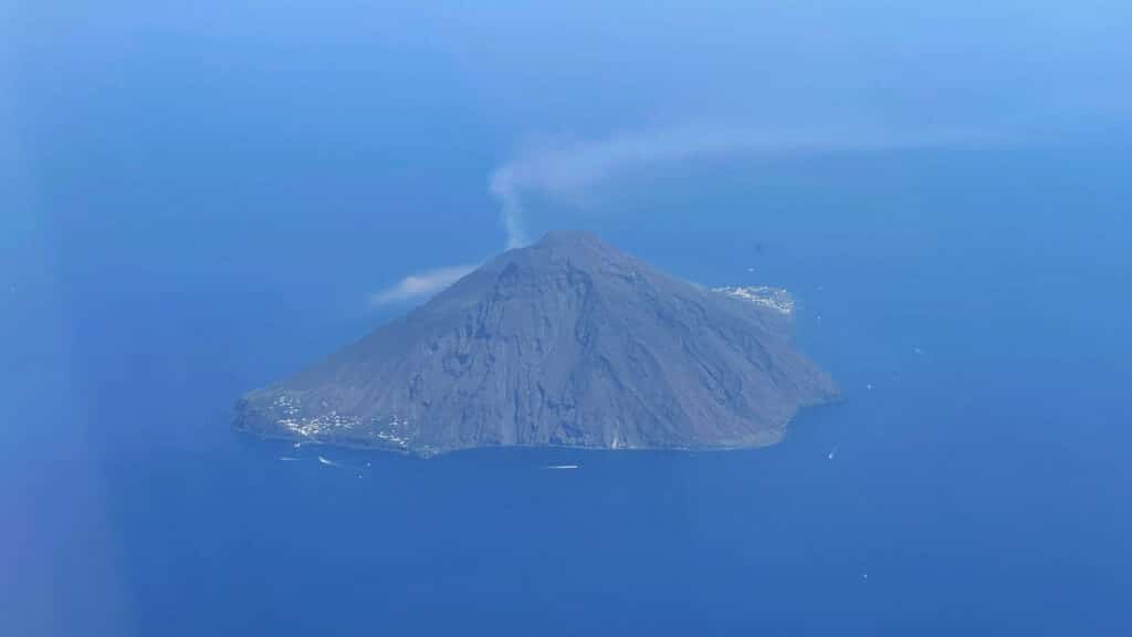 Île volcanique avec sommet actif émettant de la fumée blanche, entourée d'une mer bleue turquoise.