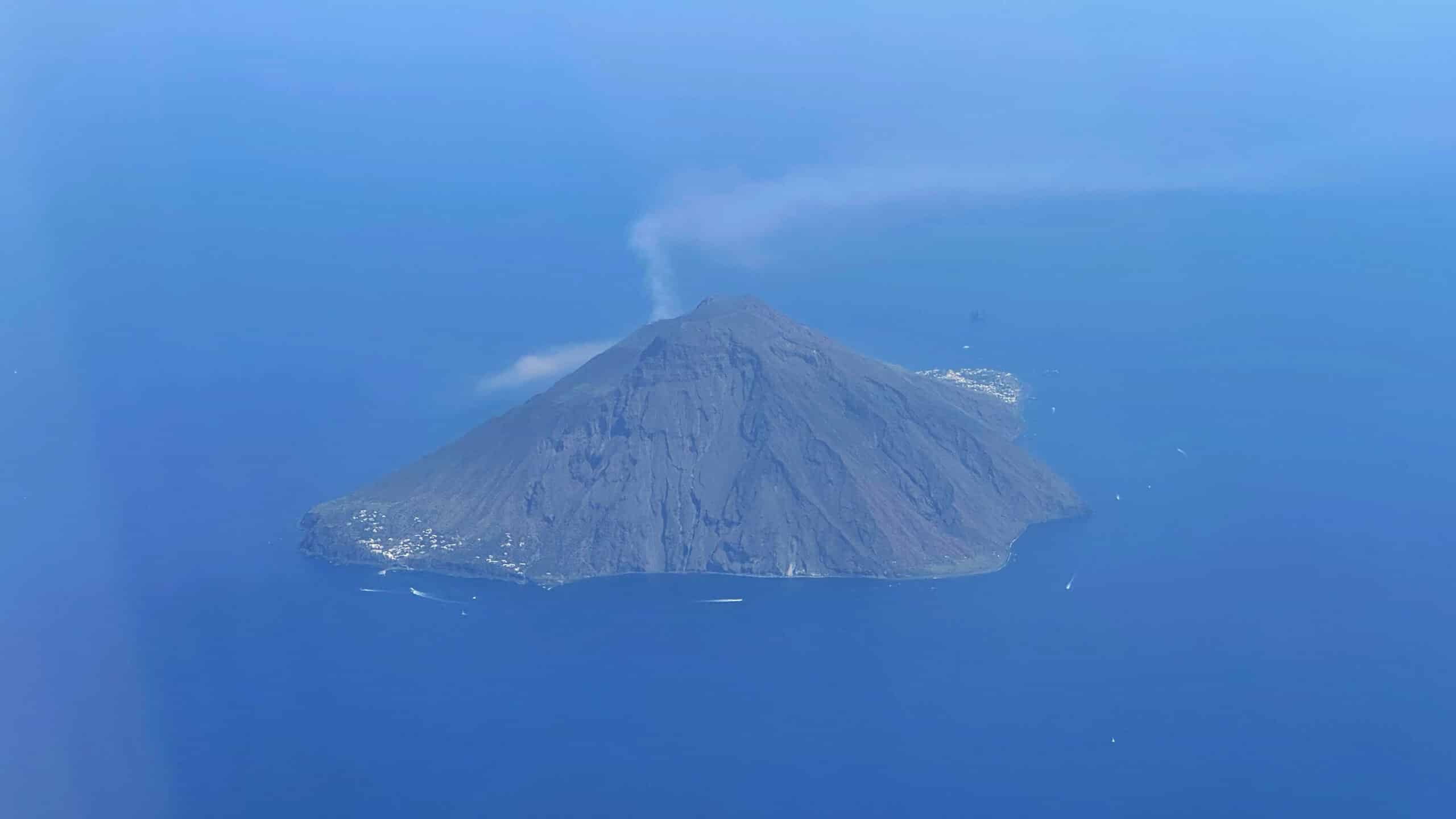 Île volcanique avec sommet actif émettant de la fumée blanche, entourée d'une mer bleue turquoise.